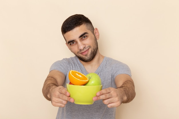 Front view young male in grey t-shirt and blue jeans holding plate with fruits smiling on beige