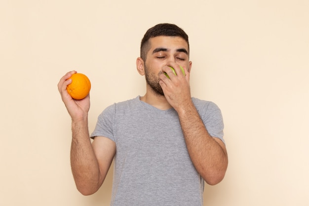 Вид спереди молодой мужчина в серой футболке и синих джинсах, пахнущий яблоком, держит апельсин на бежевом