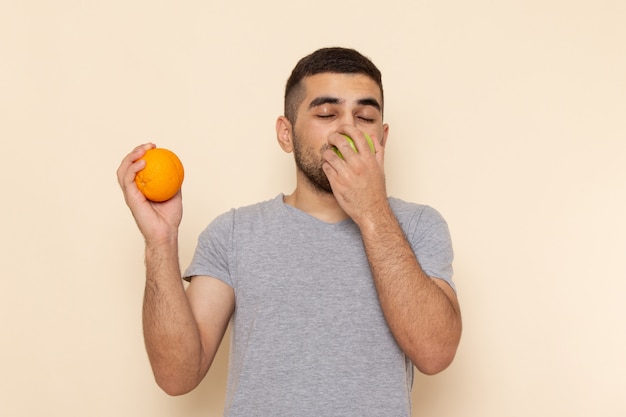 Front view young male in grey t-shirt and blue jeans smelling apple holding orange on beige