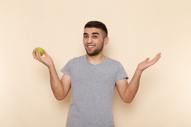 Front view young male in grey t-shirt and blue jeans smiling and holding apple on beig