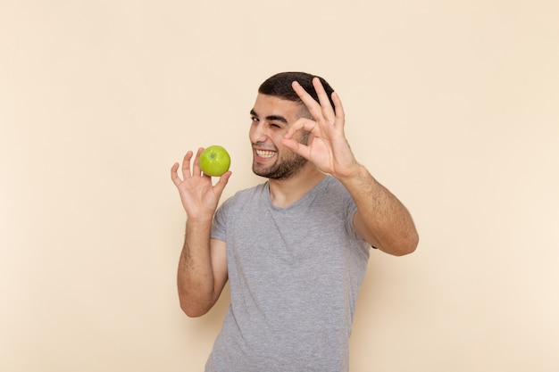 Вид спереди молодой мужчина в серой футболке и синих джинсах улыбается и держит зеленое яблоко на бежевом