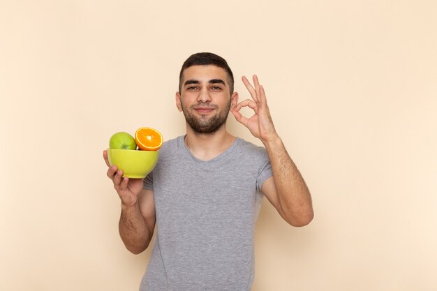 Вид спереди молодой мужчина в серой футболке и синих джинсах улыбается и держит тарелку с фруктами на бежевом