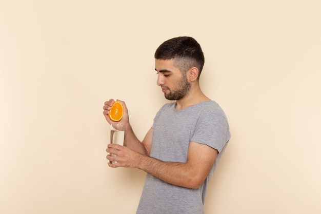 Вид спереди молодой мужчина в серой футболке и синих джинсах, выжимающий апельсиновый сок на бежевом