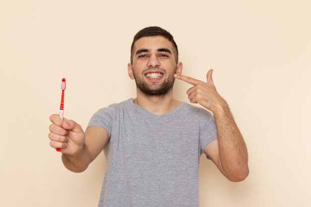 Front view young male in grey t-shirt cleaning his teeth on beige