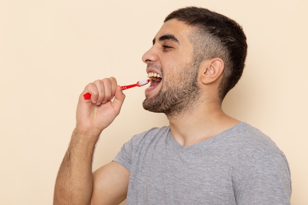 Front view young male in grey t-shirt cleaning his teeth on beige