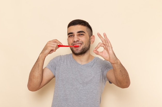Вид спереди молодой мужчина в серой футболке чистит зубы и улыбается на бежевом