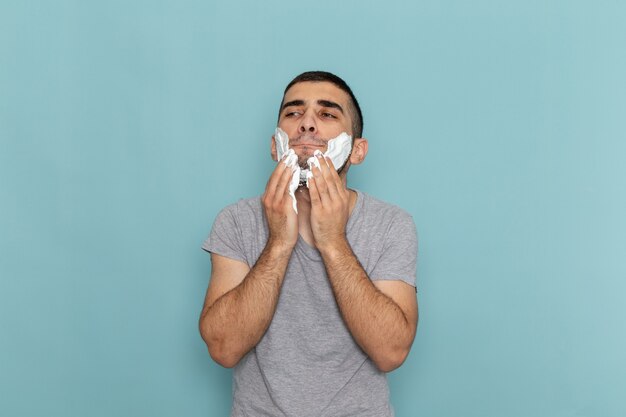 Front view young male in grey t-shirt covering his face with white foam for shaving on ice-blue wall beard foam hair razor shave