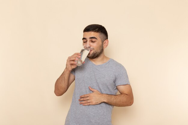 Front view young male in grey t-shirt drinking glass of water on beige