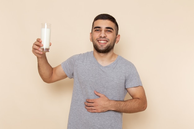 Вид спереди молодой мужчина в серой футболке пьет молоко с улыбкой на бежевом