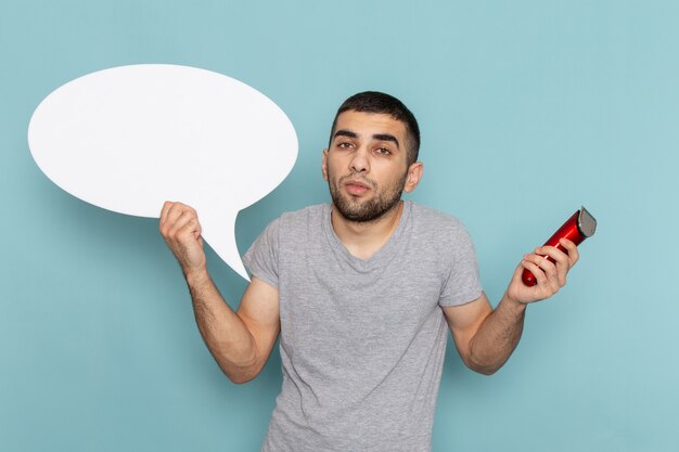 Front view young male in grey t-shirt holding a big white sign and electric razor on the ice blue wall beard hair razor shaving