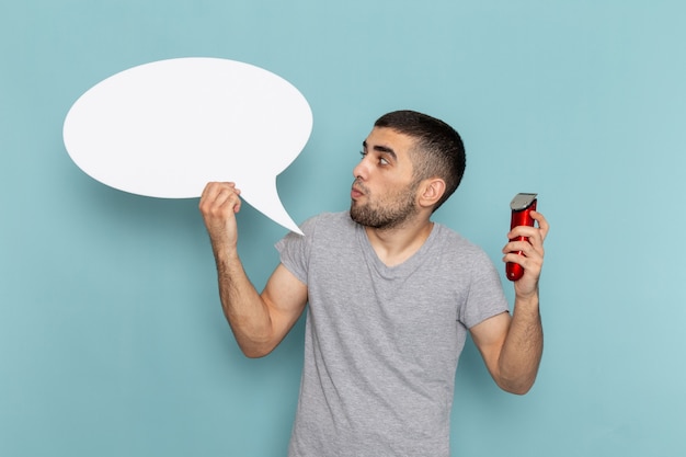 Front view young male in grey t-shirt holding a big white sign and electric razor on ice blue wall male beard hair razor shaving