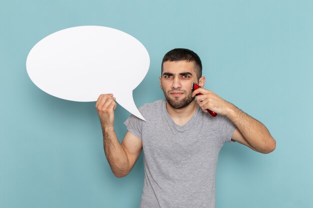 Front view young male in grey t-shirt holding big white sign and electric razor on ice blue