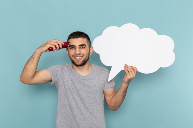 Front view young male in grey t-shirt holding a big white sign and electric razor smiling on iced blue wall male beard hair razor shaving