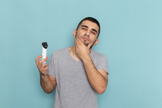 Front view young male in grey t-shirt holding electric razor on the ice-blue male shaving beard hair foam