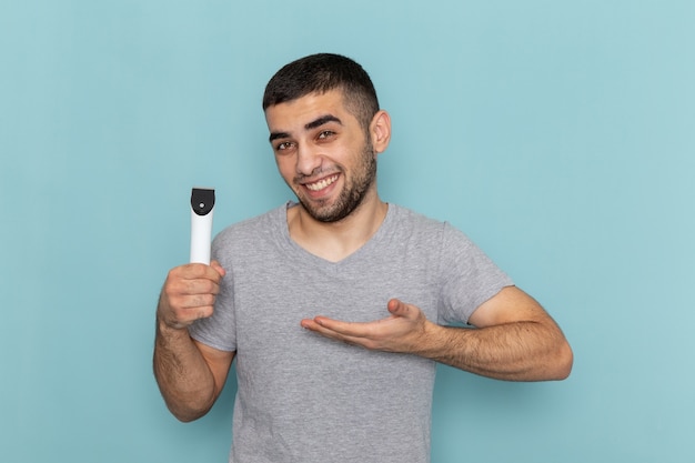 Front view young male in grey t-shirt holding electric razor with a smile on blue shaving beard male