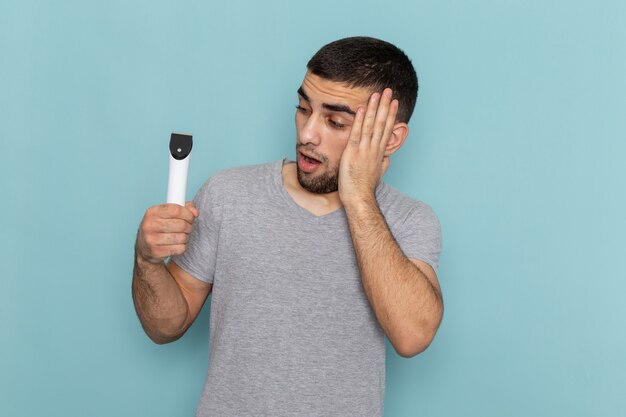 青に驚きの表情で電気かみそりを保持している灰色のTシャツの正面図若い男性