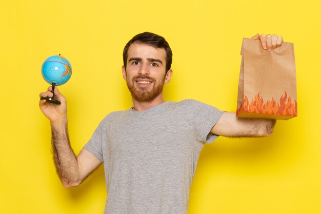 A front view young male in grey t-shirt holding food package and little globe on the yellow wall man color model emotion clothes