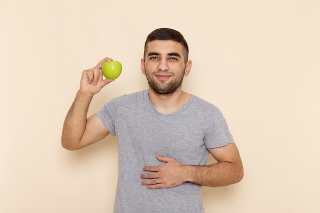 Front view young male in grey t-shirt holding green apple on beige