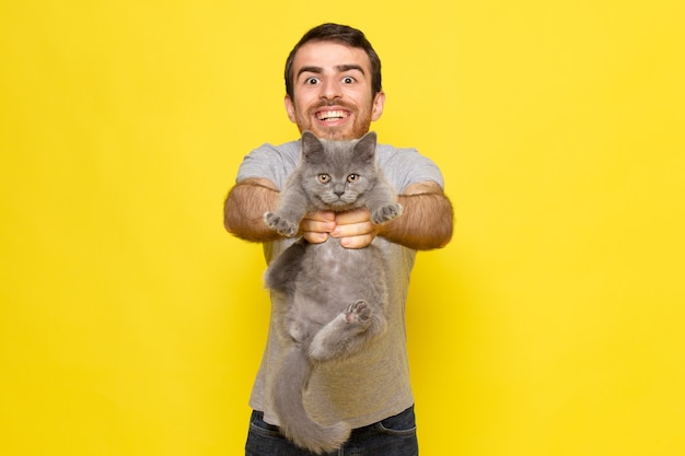 Foto gratuita un giovane maschio di vista frontale in maglietta grigia che tiene gattino grigio con il sorriso sul modello di colore di emozione di espressione dell'uomo della parete gialla