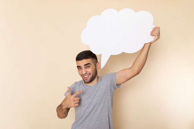 Front view young male in grey t-shirt holding huge white sign with smile on his face on beige