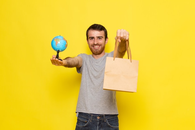 Вид спереди молодой мужчина в серой футболке, держащий маленький глобус и пакет с улыбкой на желтой стене, цветовая модель выражения эмоций человека
