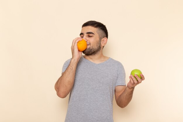 Front view young male in grey t-shirt holding orange and apple on beige