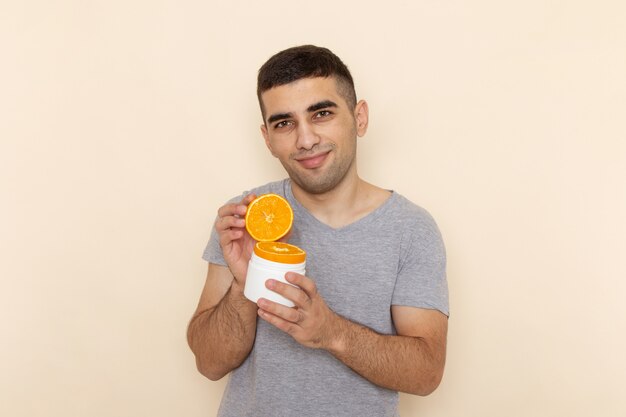 Вид спереди молодого мужчины в серой футболке, держащего дольки апельсина с легкой улыбкой на бежевом