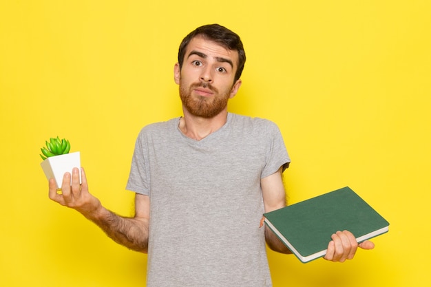 A front view young male in grey t-shirt holding plant and copybook on the yellow wall man expression emotion color model