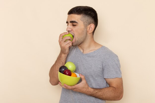 Front view young male in grey t-shirt holding plate with fruits biting apple on beige