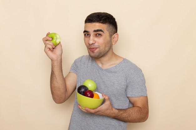 Front view young male in grey t-shirt holding plate with fruits biting apple smiling on beige