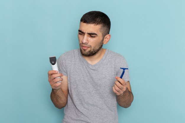 Front view young male in grey t-shirt holding razor and electric razor on blue