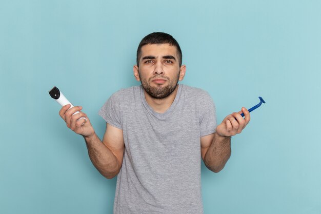 青にかみそりと電気かみそりを保持している灰色のTシャツの正面図若い男性
