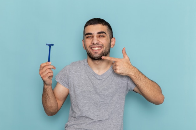 Front view young male in grey t-shirt holding razor and smiling on blue shaving beard