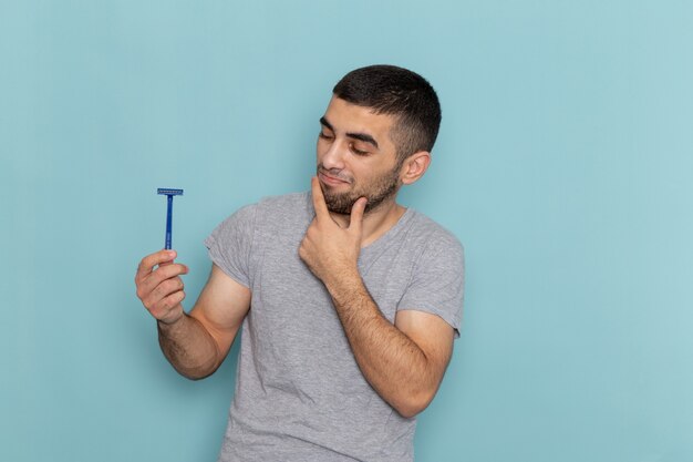 Front view young male in grey t-shirt holding razor and thinking on blue shaving beard male hair foam color