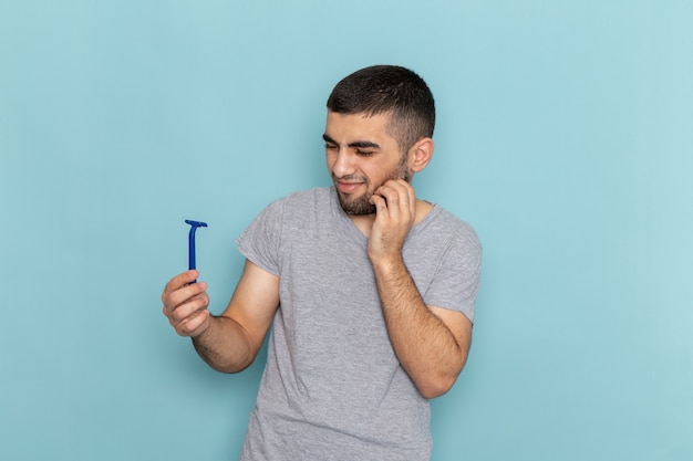 Front view young male in grey t-shirt holding razor with displeased expression on the blue