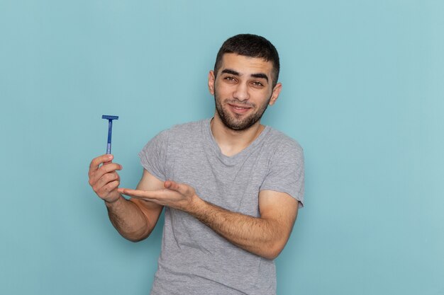 Front view young male in grey t-shirt holding razor with slight smile on blue shaving beard male hair foam color