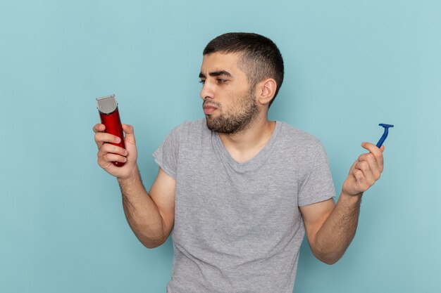 赤い電気かみそりとシンプルなかみそりをアイスブルーのひげの泡の髪かみそりのひげそりに保持している灰色のTシャツの正面図