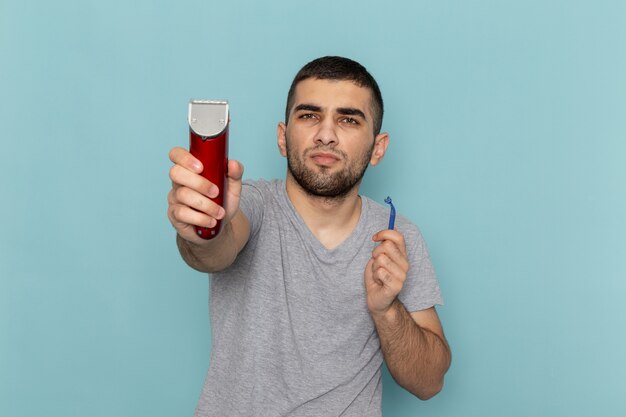 Front view young male in grey t-shirt holding red electric razor and simple razor on ice-blue beard foam razor shave