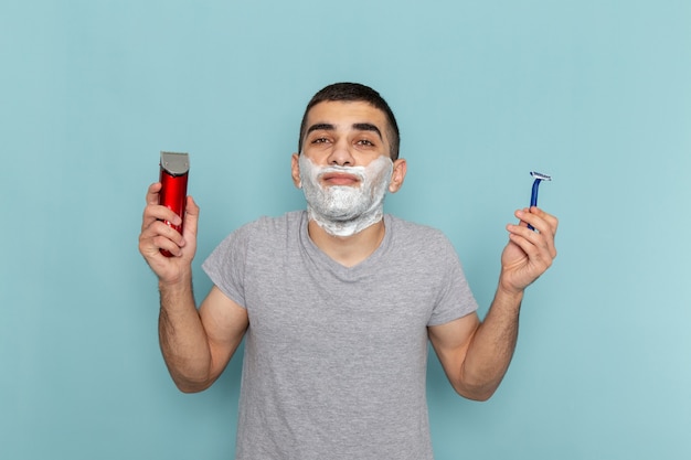 Front view young male in grey t-shirt holding simple and electric razor on ice-blue beard foam shaving male