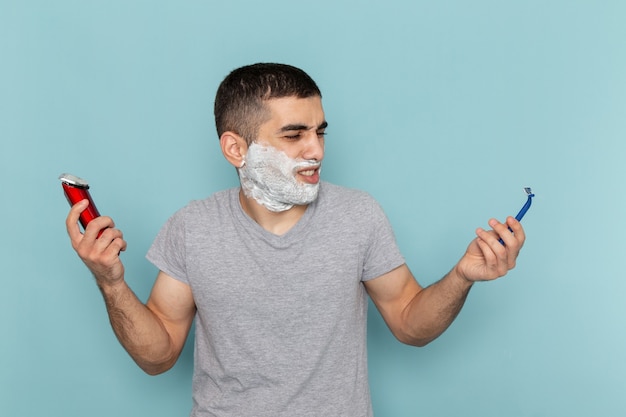 Front view young male in grey t-shirt holding simple and electric razor on iced-blue beard foam shaving male