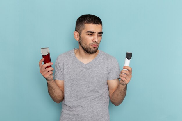 Front view young male in grey t-shirt holding two different electric razors on ice-blue male shaving beard hair foam