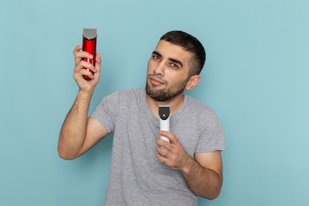 Front view young male in grey t-shirt holding two different electric razors on ice-blue