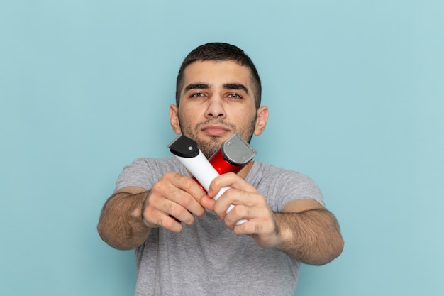 Front view young male in grey t-shirt holding two different electric razors on the ice-blue