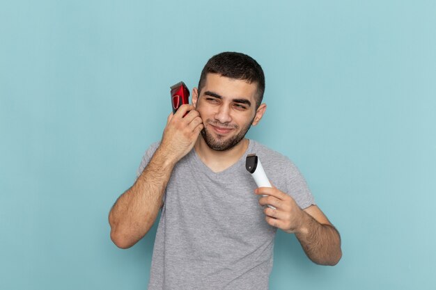 Front view young male in grey t-shirt holding two different electric razors on iced-blue