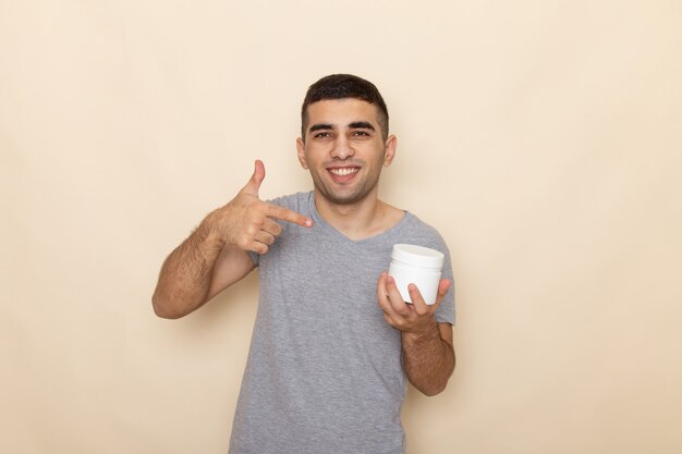 Вид спереди молодого мужчины в серой футболке, держащего белую банку с улыбкой на бежевом