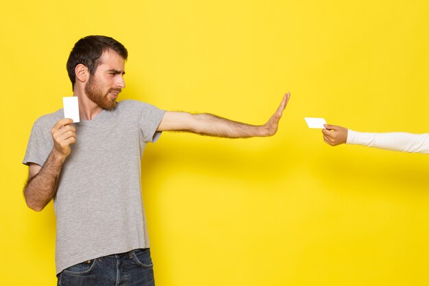 A front view young male in grey t-shirt holding white card refusing another white card on the yellow wall man color model emotion clothes