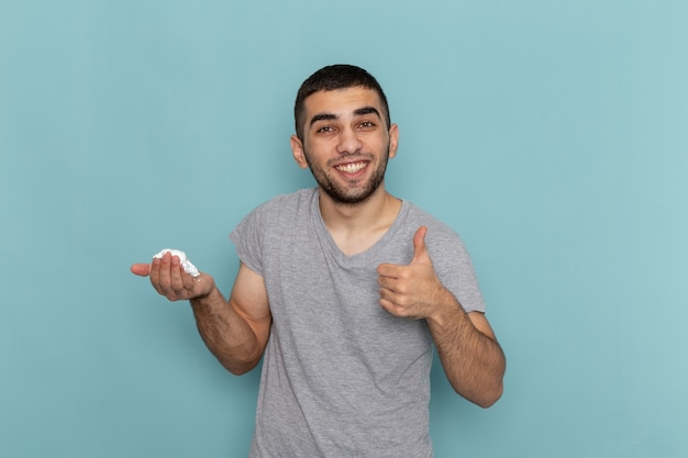 Front view young male in grey t-shirt holding white foam for shaving smiling on ice-blue