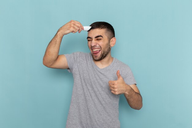 Front view young male in grey t-shirt shaving his head with electric razor on ice-blue