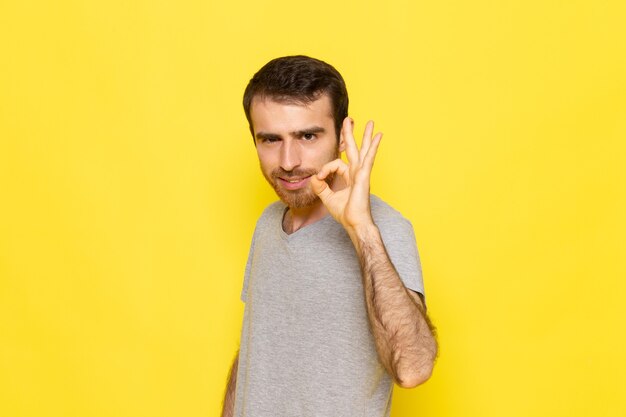 Молодой мужчина в серой футболке, вид спереди, показывает знак на желтой стене, цветная модель, эмоция, одежда