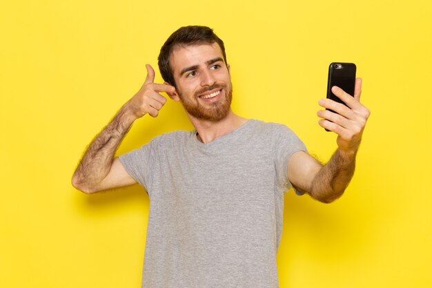 Молодой мужчина в серой футболке улыбается и делает селфи на желтой стене, цветная модель, эмоция, одежда